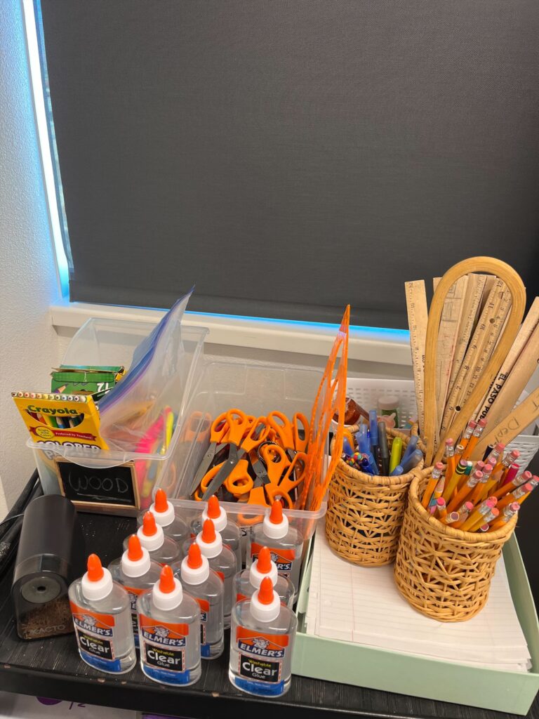 Classroom supply station with glue bottles, scissors, markers, pencils, rulers, and loose-leaf paper organized in baskets and bins.
