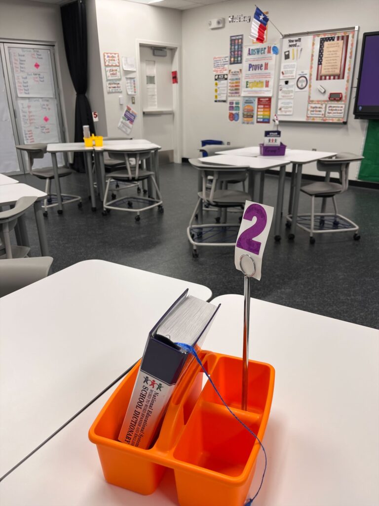 ESL classroom with grouped desks, a numbered table caddy, and visible anchor charts on writing strategies and classroom expectations.