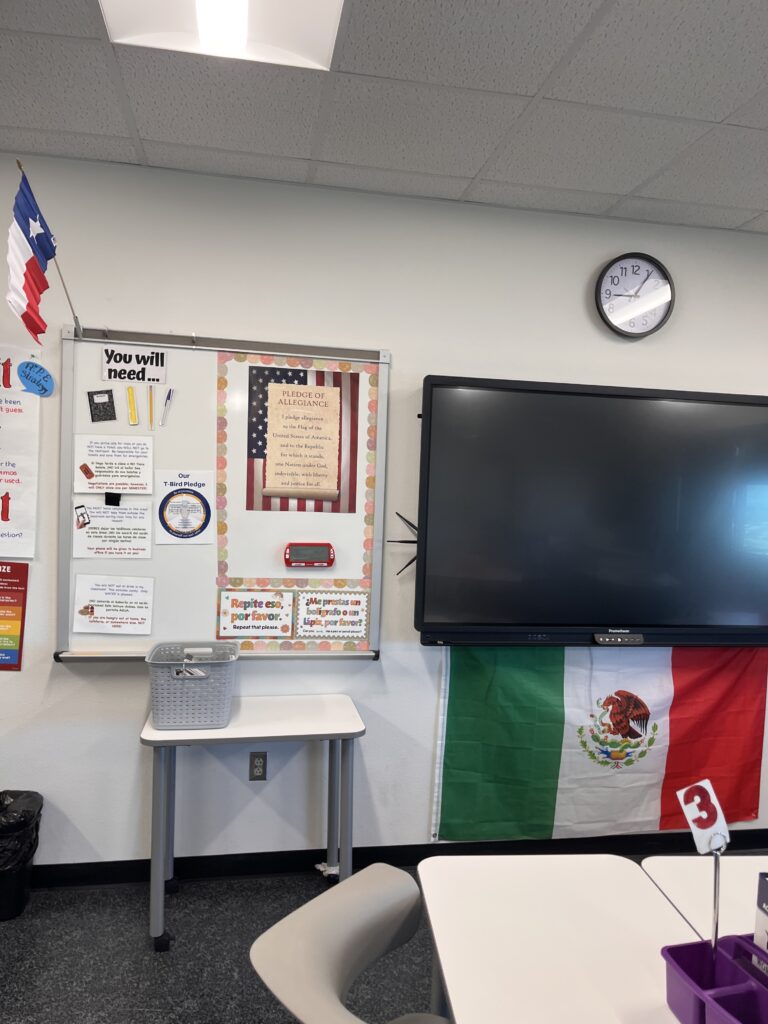 Front of an ESL classroom with digital screen, classroom supply list, the Pledge of Allegiance, and U.S. and Mexican flags.