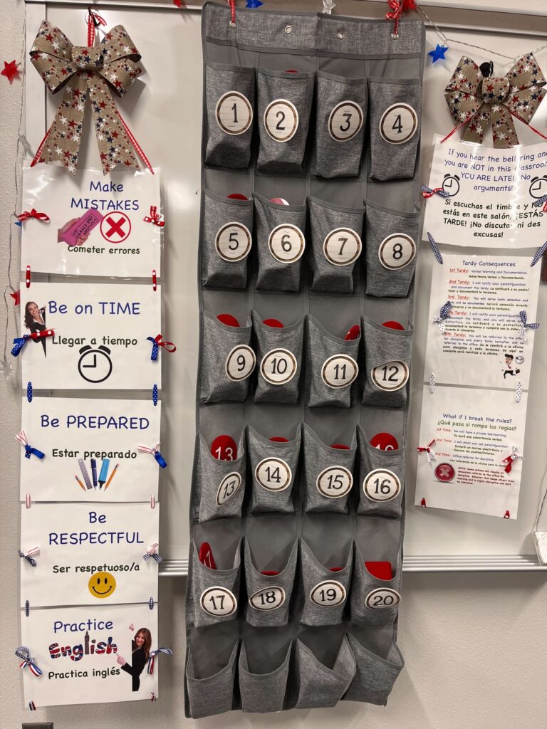 Phone storage station in an ESL classroom with numbered pockets, bilingual expectations posters, and tardy policy signs.