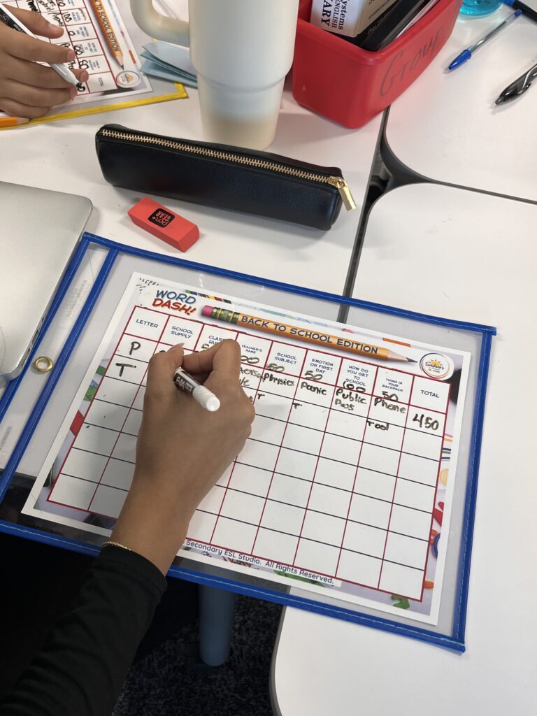 High school ESL student writing answers on a laminated Word Dash! Back to School game board during class